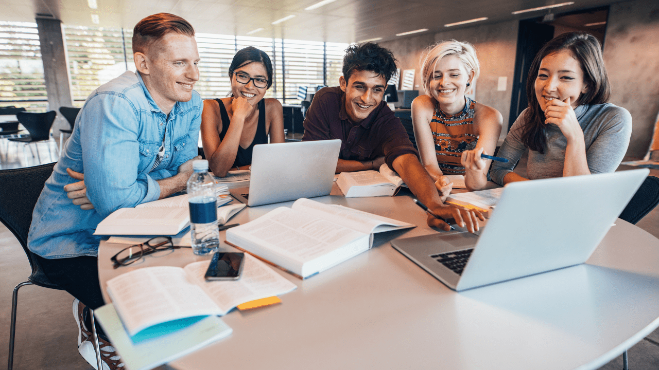 Five young entrepreneurs gathered around a table with two laptops open, discussing which software would be best for their startup business. They are dressed in casual attire and are happily engaged in an intense debate about the pros and cons of each option.