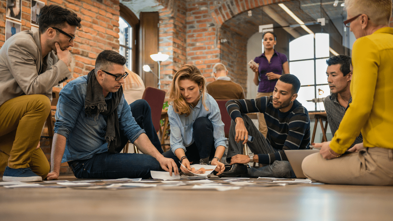 A group of diverse entrepreneurs sitting on a floor in a circle discussing business ideas, looking at notes, and sharing creative solutions.